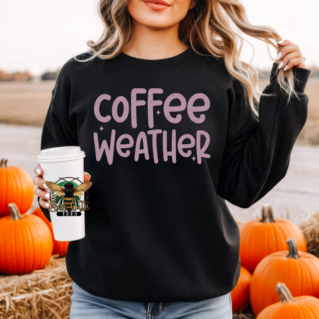 A woman wearing a black crewneck sweatshirt with 'coffee weather' printed in pink on the front, holding a white coffee cup with a bee logo. She is standing outdoors among pumpkins and hay.