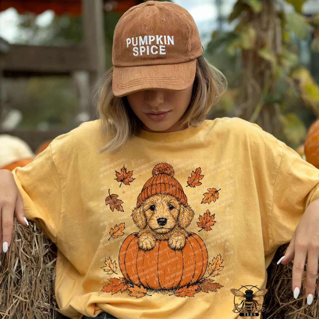 A woman wearing a tan baseball cap that says 'PUMPKIN SPICE' and a yellow graphic t-shirt featuring a puppy in a knit hat sitting on a pumpkin, posed on hay bales at what appears to be an autumn farm setting.
