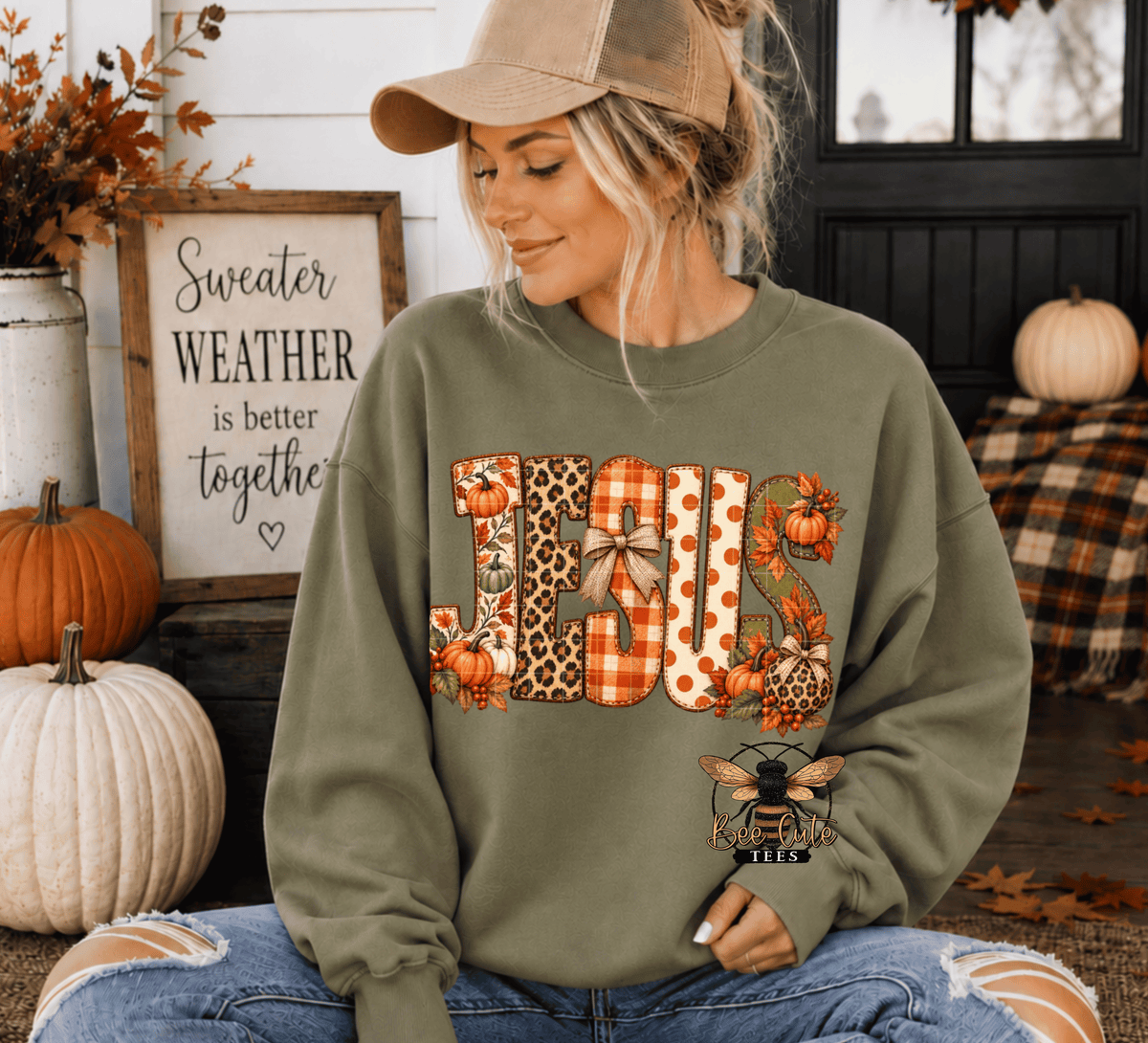 A woman wearing an olive green crewneck sweatshirt with a large 'JESUS' fall-themed graphic and a bee patch, sitting in a cozy autumn porch setting with pumpkins and fall decorations.