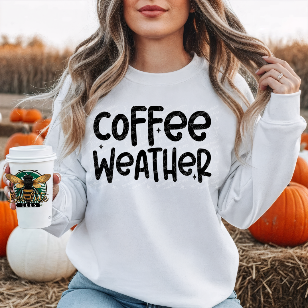 A woman in a white sweatshirt with 'coffee weather' printed in black text, holding a  coffee cup in a pumpkin patch setting.