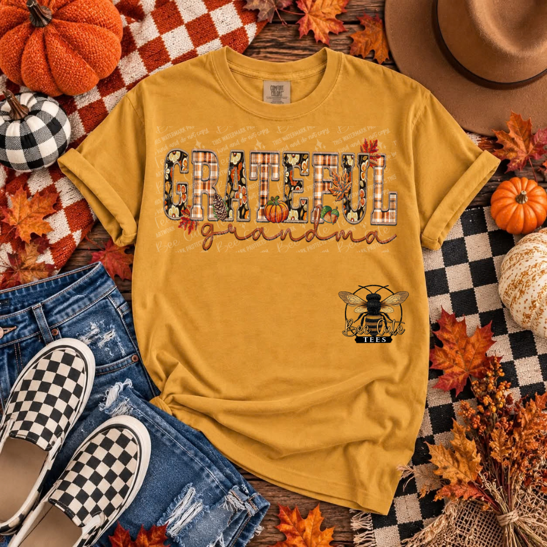 A mustard yellow graphic t-shirt laid flat in a cozy autumn-themed flat lay. The shirt features the text 'GRATEFUL Grandma' in a decorative block font with fall leaves and pumpkins, and a small bee logo underneath.