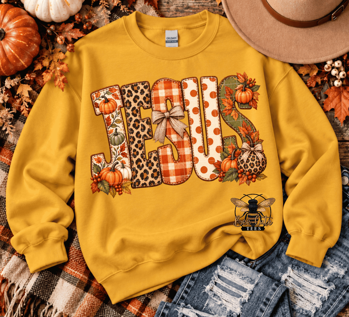 A yellow crewneck sweatshirt laid flat with a fall-themed graphic spelling 'JESUS' in patterned letters, surrounded by pumpkins, leaves, a hat, and denim.