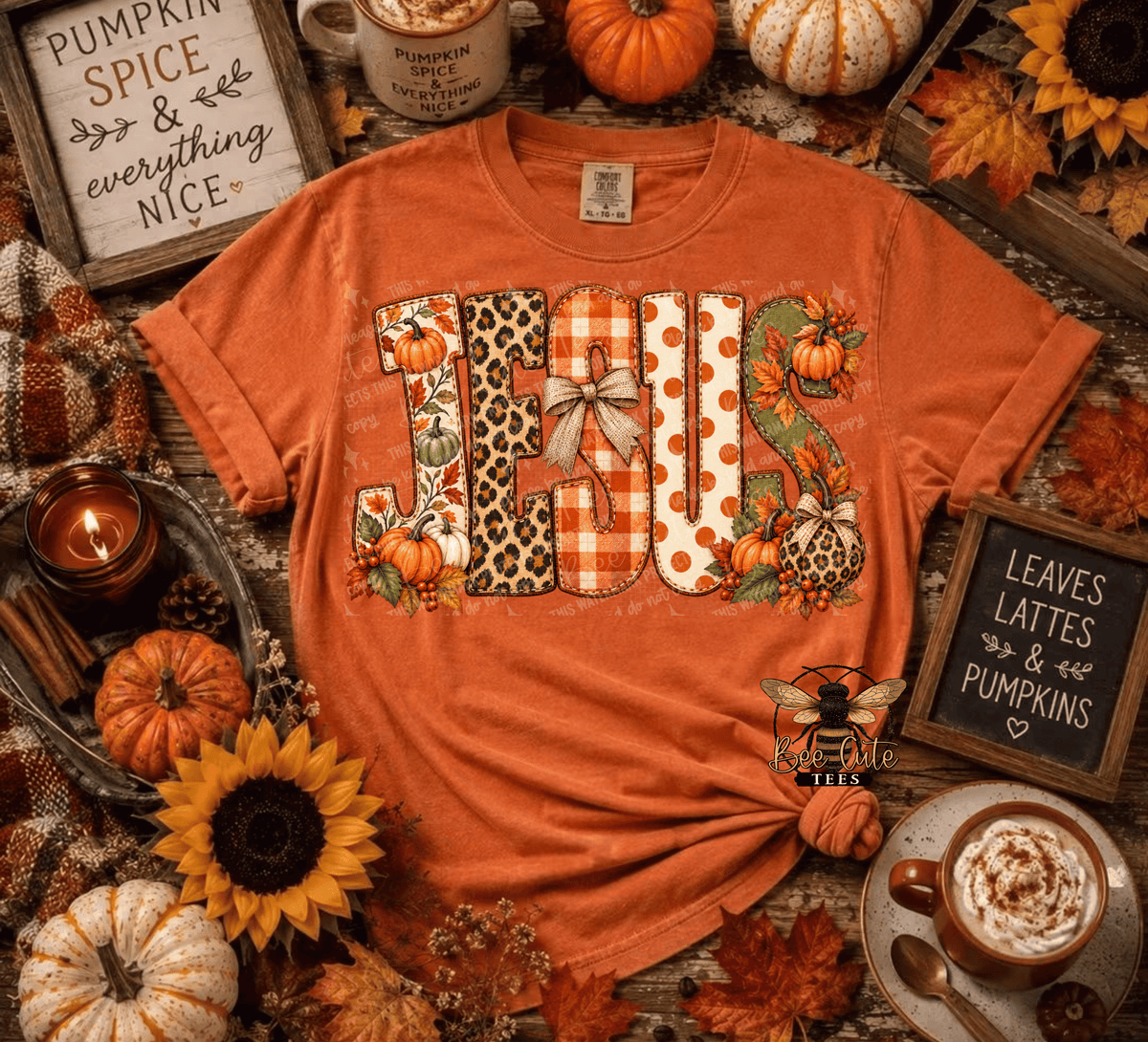 An orange graphic t-shirt featuring the word 'JESUS' decorated with autumn patterns like leopard print, gingham, polka dots, and pumpkins. The shirt is styled in a cozy fall flat-lay with pumpkins, sunflowers, a hot cocoa cup, and autumn signs.