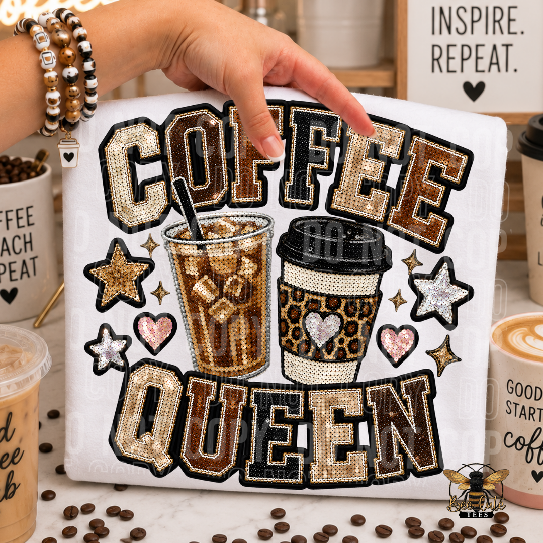 A white folded t-shirt featuring the words 'COFFEE QUEEN' in large outlined letters, with two illustrated coffee cups and decorative stars and hearts. A hand is pointing at the design, and the item is photographed in a styled kitchen-like setting with coffee cups, beans, and sign decor around it.