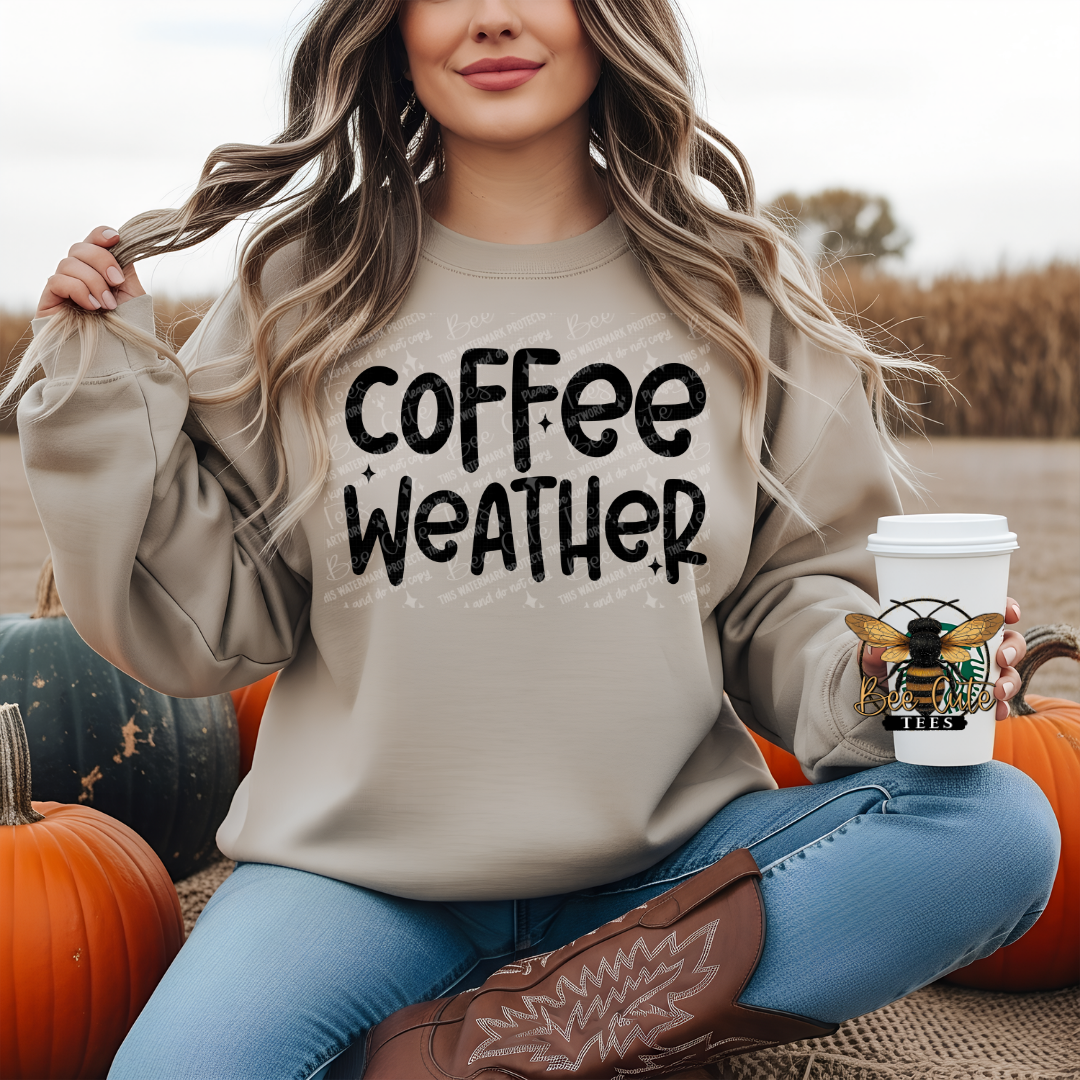 A woman sitting outdoors among pumpkins wears a beige crewneck sweatshirt with the phrase 'coffee weather' printed in black across the front. She holds a white coffee cup with a bee logo and a boot in a fall-themed setting.
