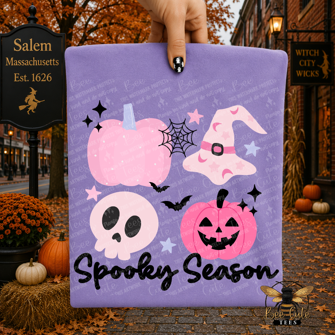 A hand holds a purple folded t-shirt or card featuring a Halloween graphic with pink pumpkins, a witch hat, a skull, bats, stars, and the text 'Spooky Season.' The background is a blurred street scene with autumn leaves, pumpkins, and storefront signs.