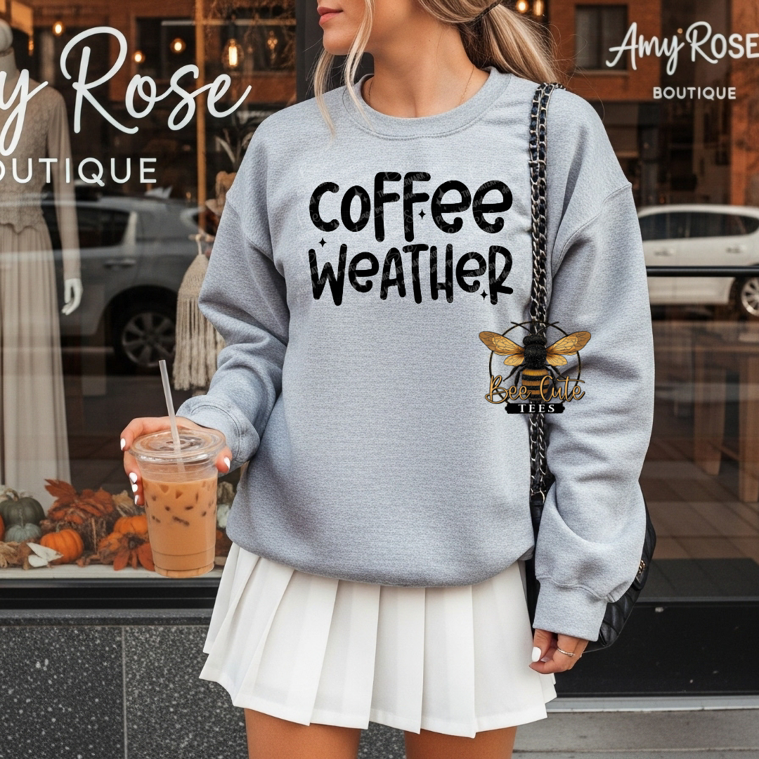 A woman wearing a light heather gray sweatshirt with the text 'coffee weather' in black, paired with a white pleated mini skirt. She is holding an iced coffee in front of a boutique window, with a black shoulder bag featuring a bee logo.