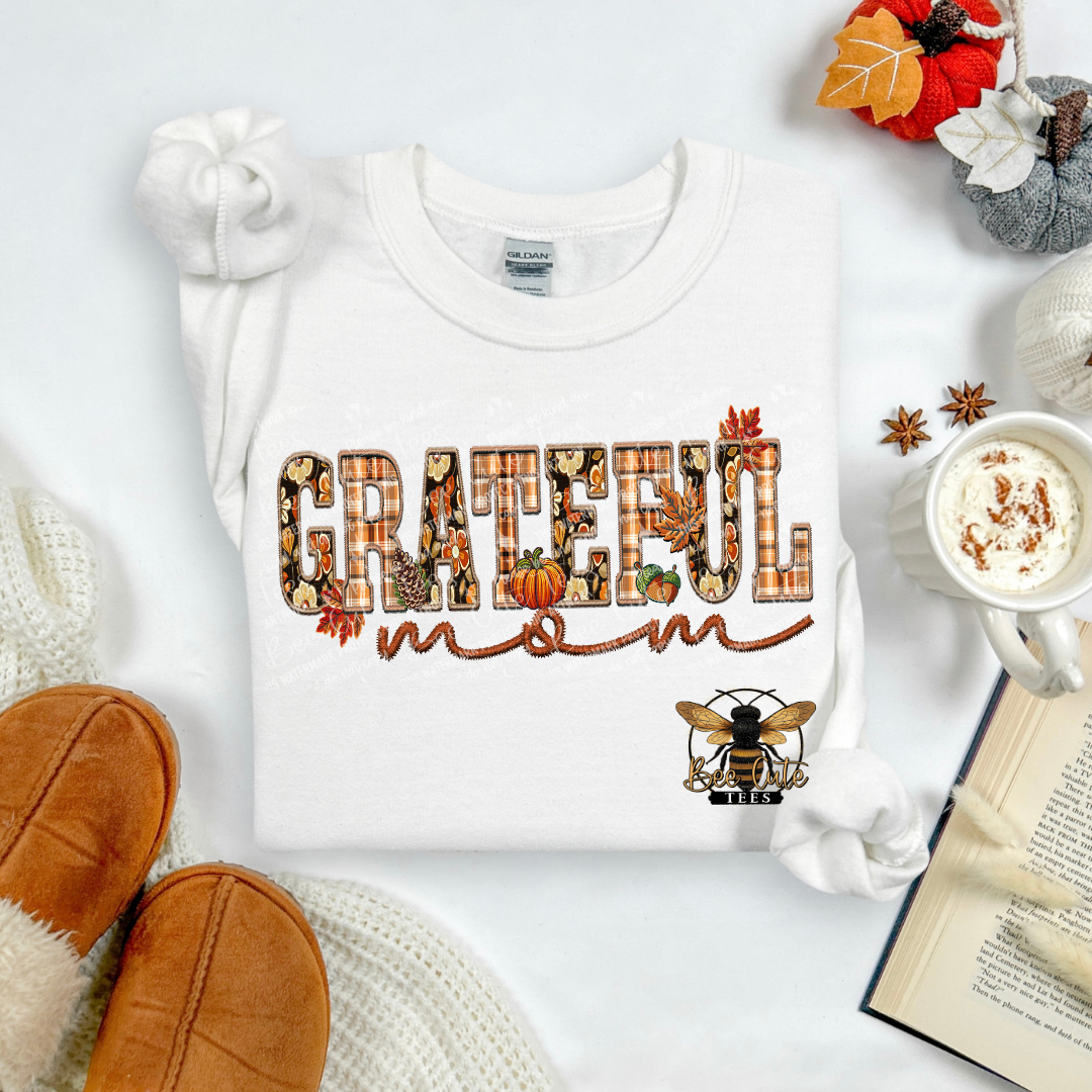 A folded white sweatshirt with a 'Grateful Mom' graphic in autumn-themed lettering, styled in a cozy flat lay with a mug of coffee, knit socks, a book, and fall leaves.