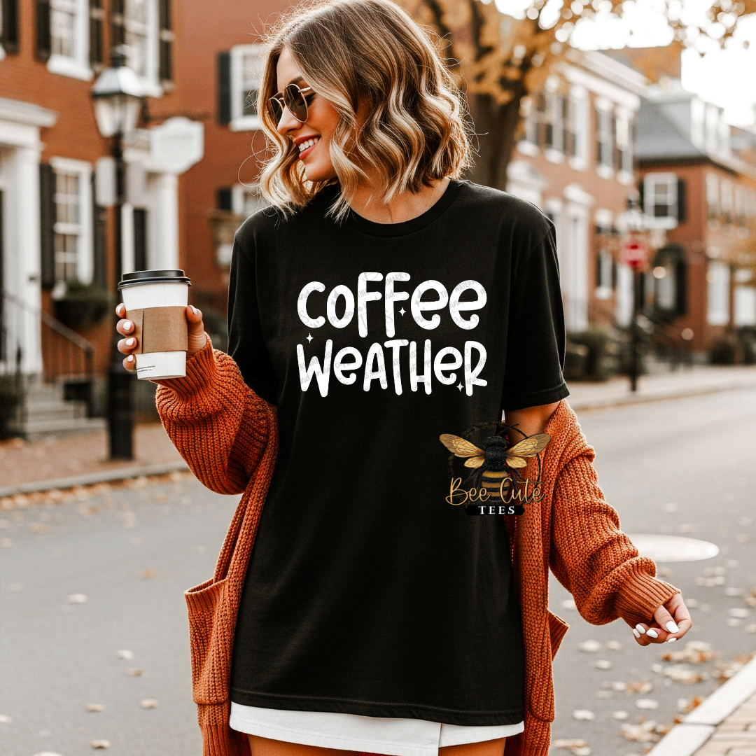 A woman wearing a black graphic t-shirt with 'coffee weather' printed in white, styled with an orange knit cardigan. She is holding a coffee cup on a city street with brick buildings in the background.