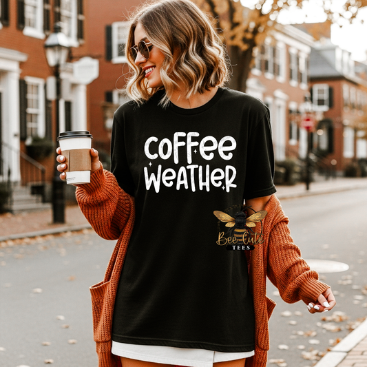 A woman wearing a black graphic t-shirt with 'coffee weather' printed in white, styled with an orange knit cardigan. She is holding a coffee cup on a city street with brick buildings in the background.