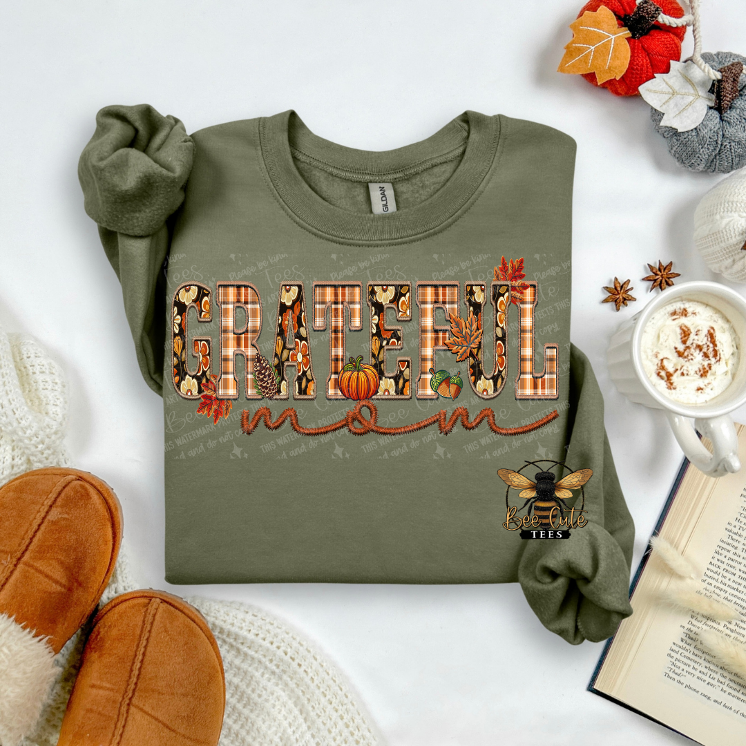 An olive green long-sleeve sweatshirt laid flat on a white surface, decorated with a 'Grateful Mom' autumn graphic. The sweatshirt is styled in a cozy flat-lay arrangement with brown suede slippers, a white knit blanket, an open book, a mug of coffee with cinnamon, and fall-themed decorations.