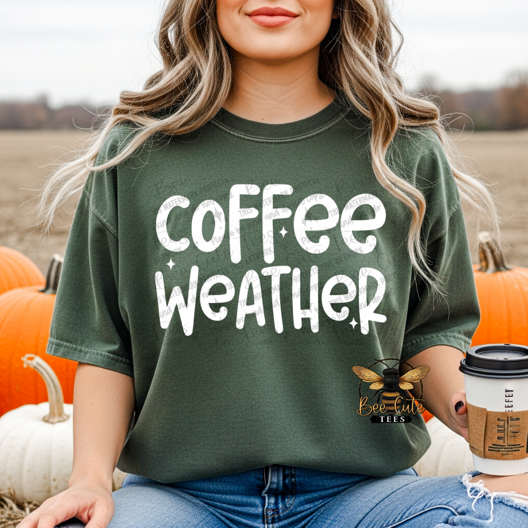 A woman wearing an oversized dark green t-shirt with 'coffee weather' printed in white distressed lettering, sitting in a pumpkin patch and holding a coffee cup.