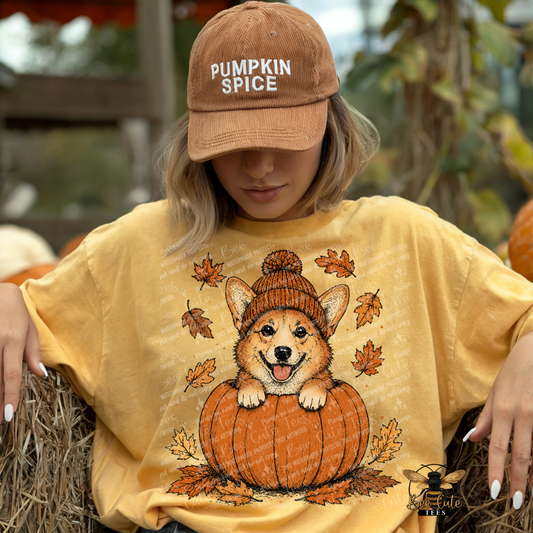 A woman wearing a tan 'Pumpkin Spice' baseball cap and a yellow graphic t-shirt featuring a corgi in a knit hat sitting on a pumpkin, posed outdoors on a hay bale with pumpkins in the background.