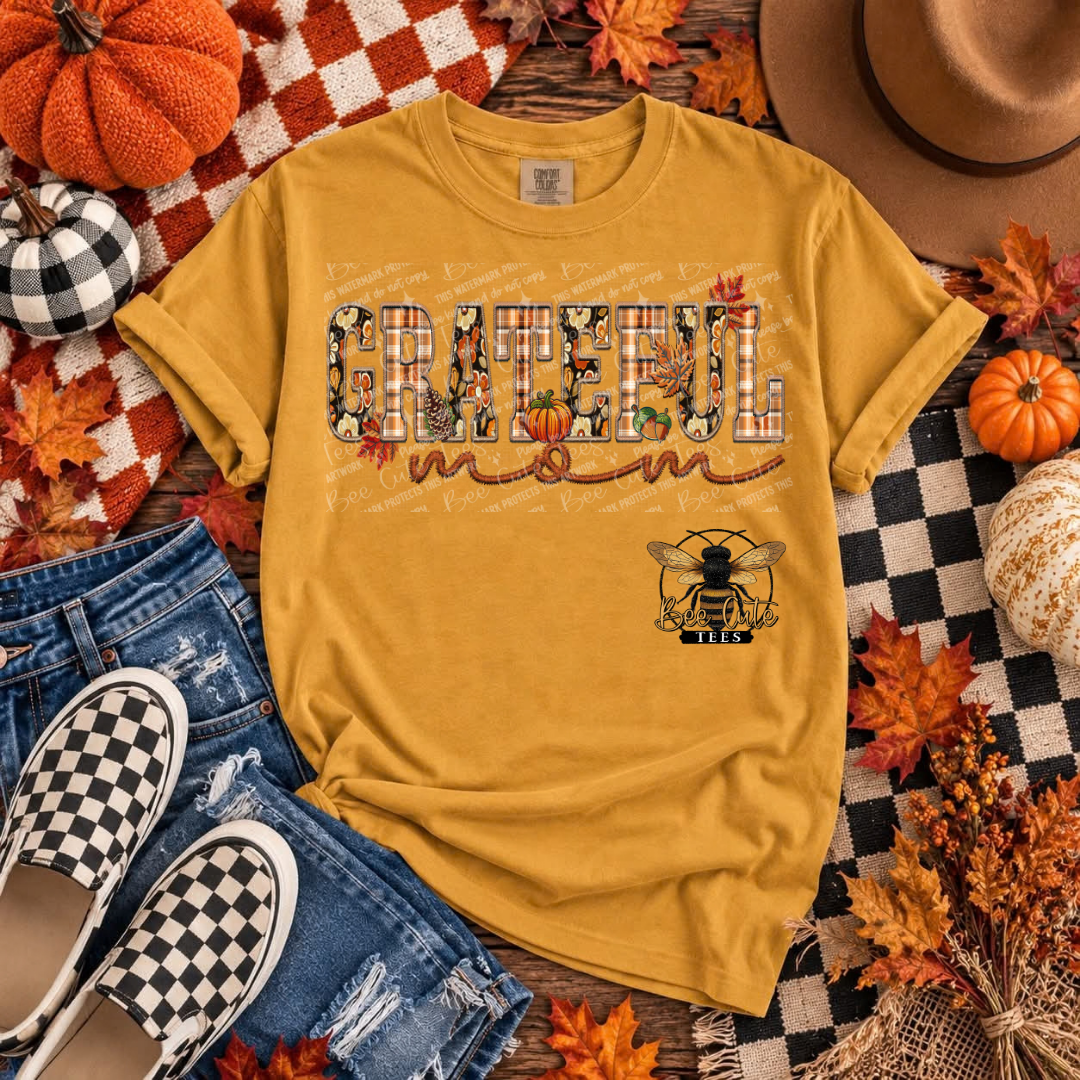 A mustard yellow graphic t-shirt laid flat in an autumn-themed flat lay with jeans, slip-on shoes, pumpkins, leaves, a hat, and a checkered blanket. The shirt features a large 'GRATEFUL mom' text design with fall icons inside the letters and a bee logo with 'GRATEFUL' on the chest.