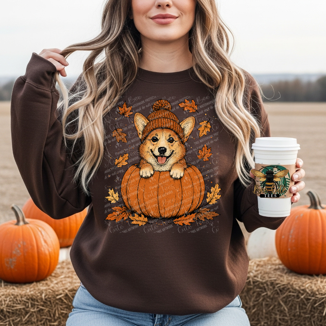 A woman wearing a brown crewneck sweatshirt with a graphic of a corgi dog sitting in a pumpkin, holding a white coffee cup. She is standing outdoors in a field with pumpkins around her.