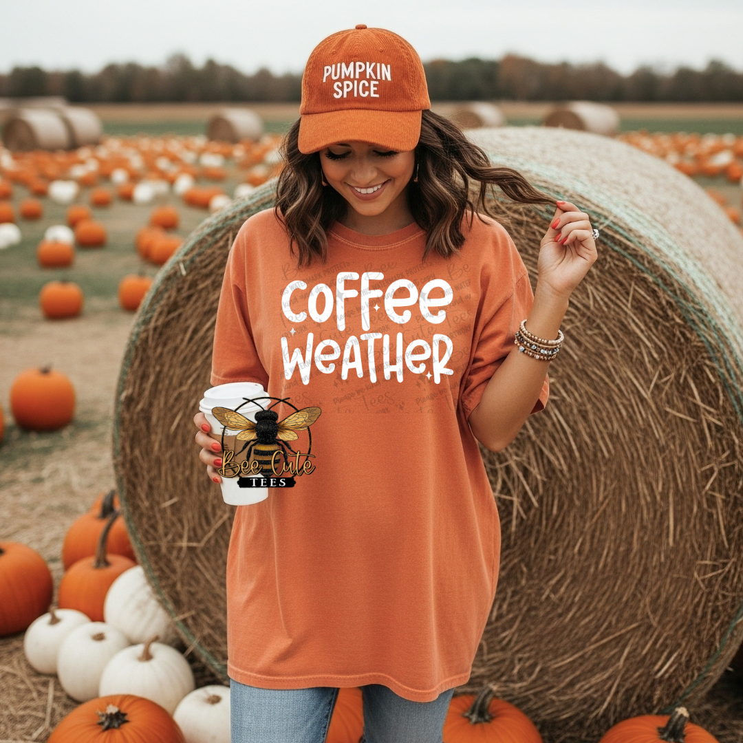 A woman standing in a pumpkin field wears an oversized burnt orange t-shirt with 'coffee weather' printed in white, paired with a matching burnt orange 'PUMPKIN SPICE' baseball cap and holding a coffee cup.