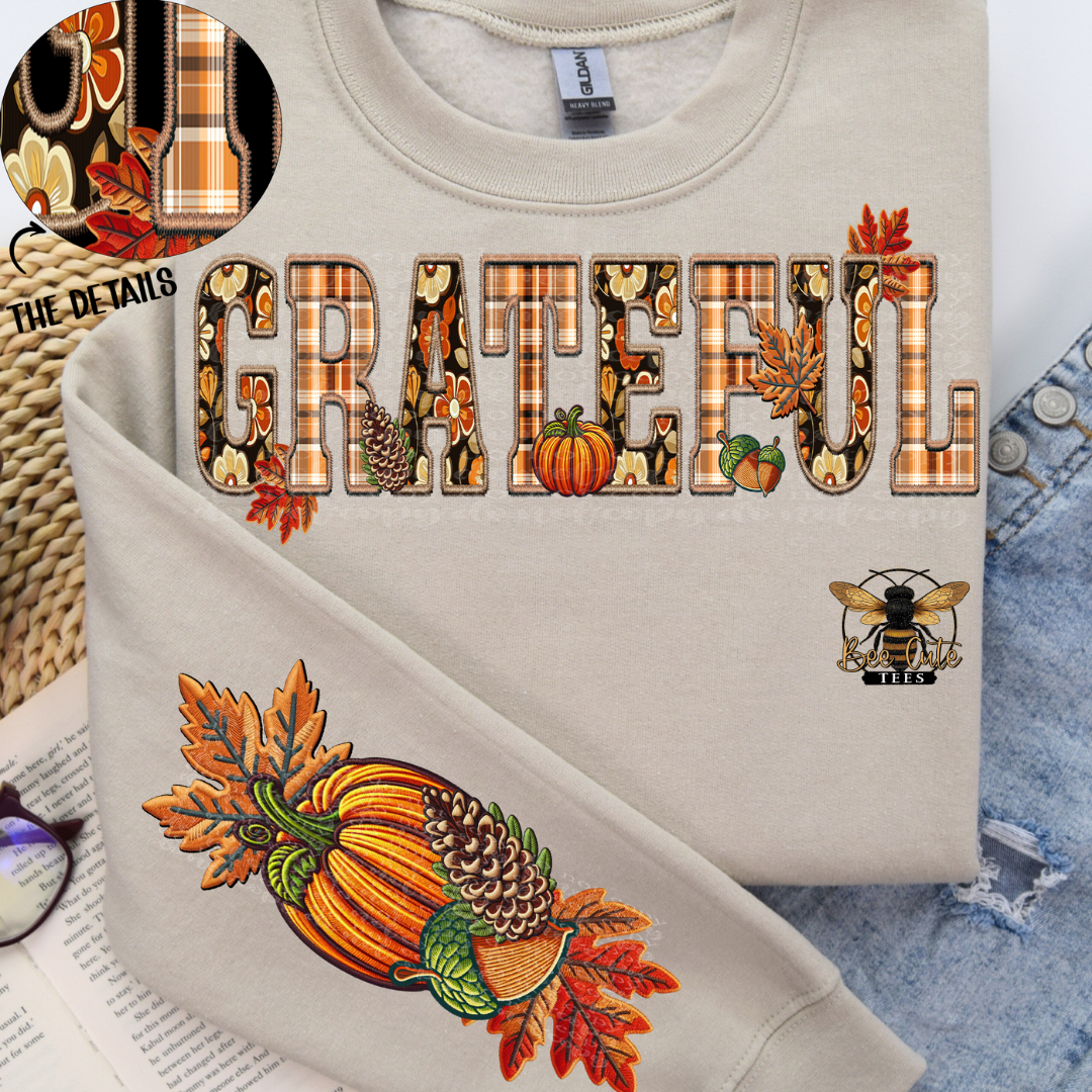A folded light beige crewneck sweatshirt with a fall-themed graphic reading 'GRATEFUL' in plaid and autumn leaf letters, plus a pumpkin and acorn illustration on the sleeve. The sweatshirt is styled on top of denim fabric with a small bee patch logo and a circular detail inset showing the print texture.