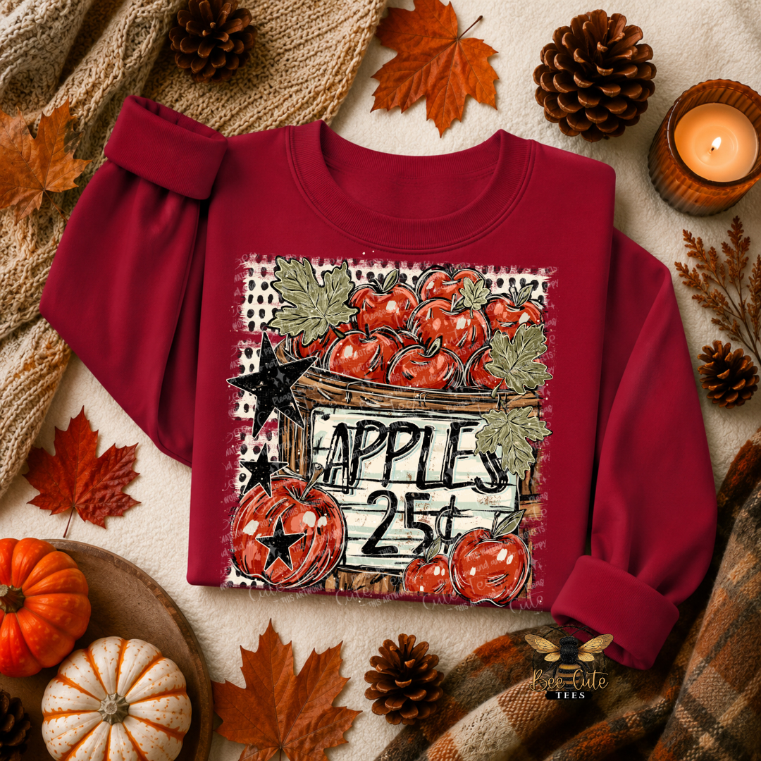 A burgundy crewneck sweatshirt with a rustic autumn-themed apple graphic on the front, styled in a cozy flat-lay with fall leaves, pumpkins, a candle, and a knitted blanket.