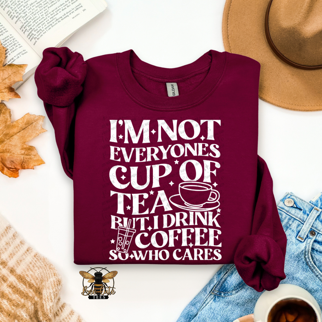 A folded maroon long-sleeve graphic T-shirt with rolled sleeves, featuring the white text 'I'M NOT EVERYONE'S CUP OF TEA BUT I DRINK COFFEE SO WHO CARES' with a coffee cup illustration. The shirt is styled in a flat lay with a tan hat, blue jeans, a book, autumn leaves, a knit sweater, and a small coffee cup on a white background.
