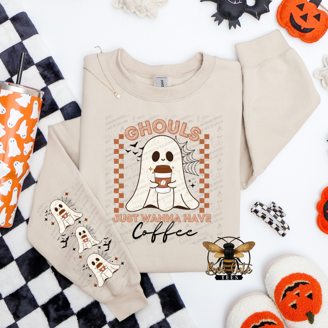 A beige long-sleeve sweatshirt with a Halloween graphic featuring a ghost holding a coffee cup and the text 'Ghouls just wanna have Coffee'. The sweatshirt is styled in a flat lay with a black and white checkered blanket, an orange cup with ghost patterns, and pumpkin decorations.