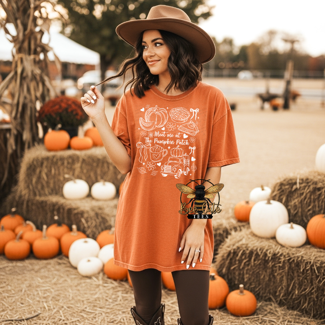 A woman wearing a brown wide-brim hat and a rust-orange graphic t-shirt stands in a fall-themed outdoor setting with pumpkins and hay bales. The t-shirt features a white line art graphic of autumn foods and the text 'Meet me at The Pumpkin Patch'.