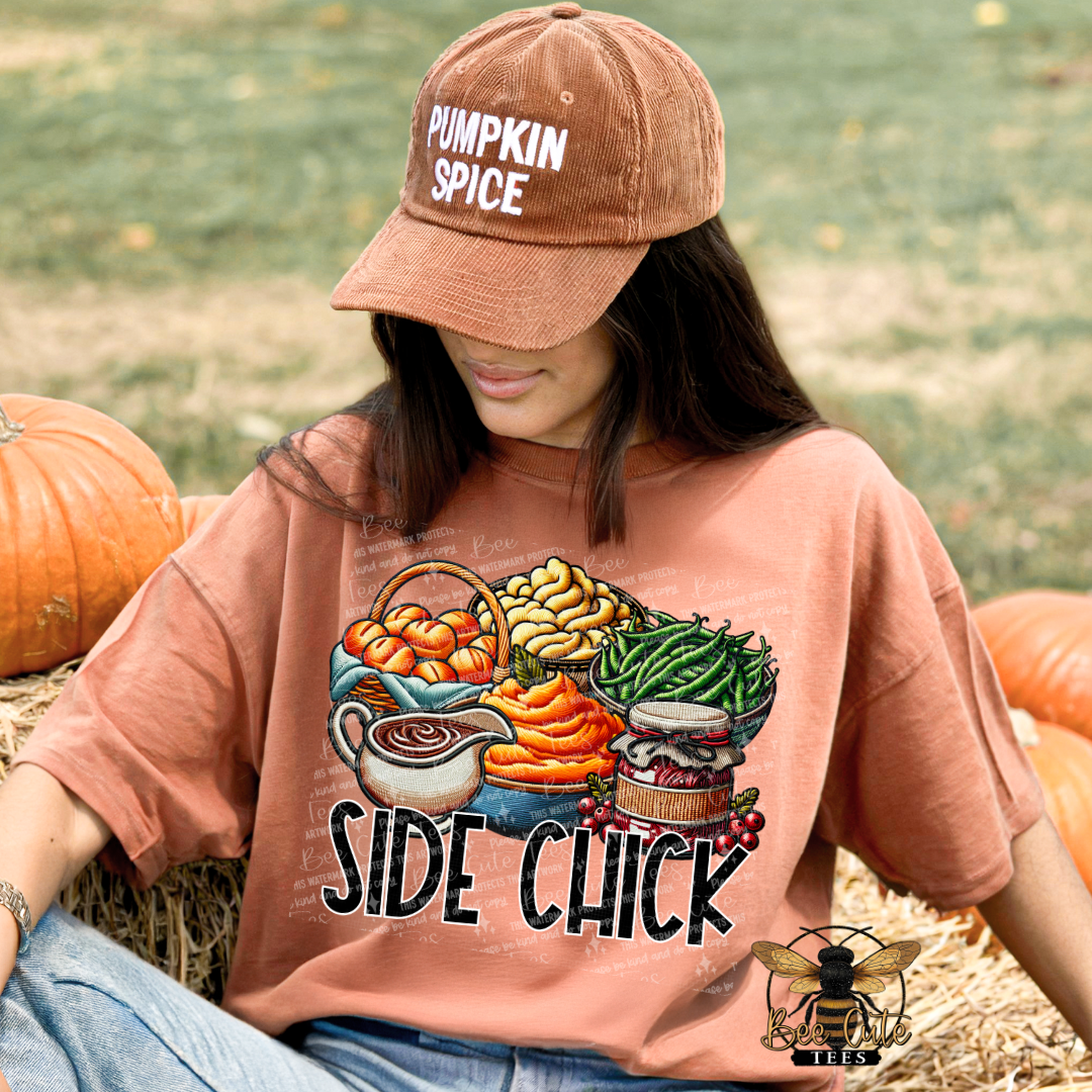 A woman wearing a dusty rose graphic t-shirt and a brown baseball cap with 'PUMPKIN SPICE' written on it, sitting on a hay bale with pumpkins in the background. The t-shirt features a colorful autumn harvest graphic with the text 'SIDE CHICK'.