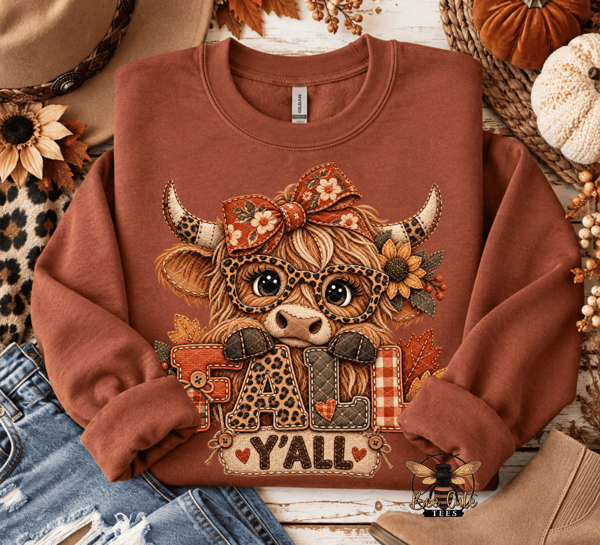 A rust-brown crewneck sweatshirt laid flat with autumn-themed props. The front has a large graphic of a highland cow wearing a bow and glasses, with the word 'FALL Y'all' in decorative letters.