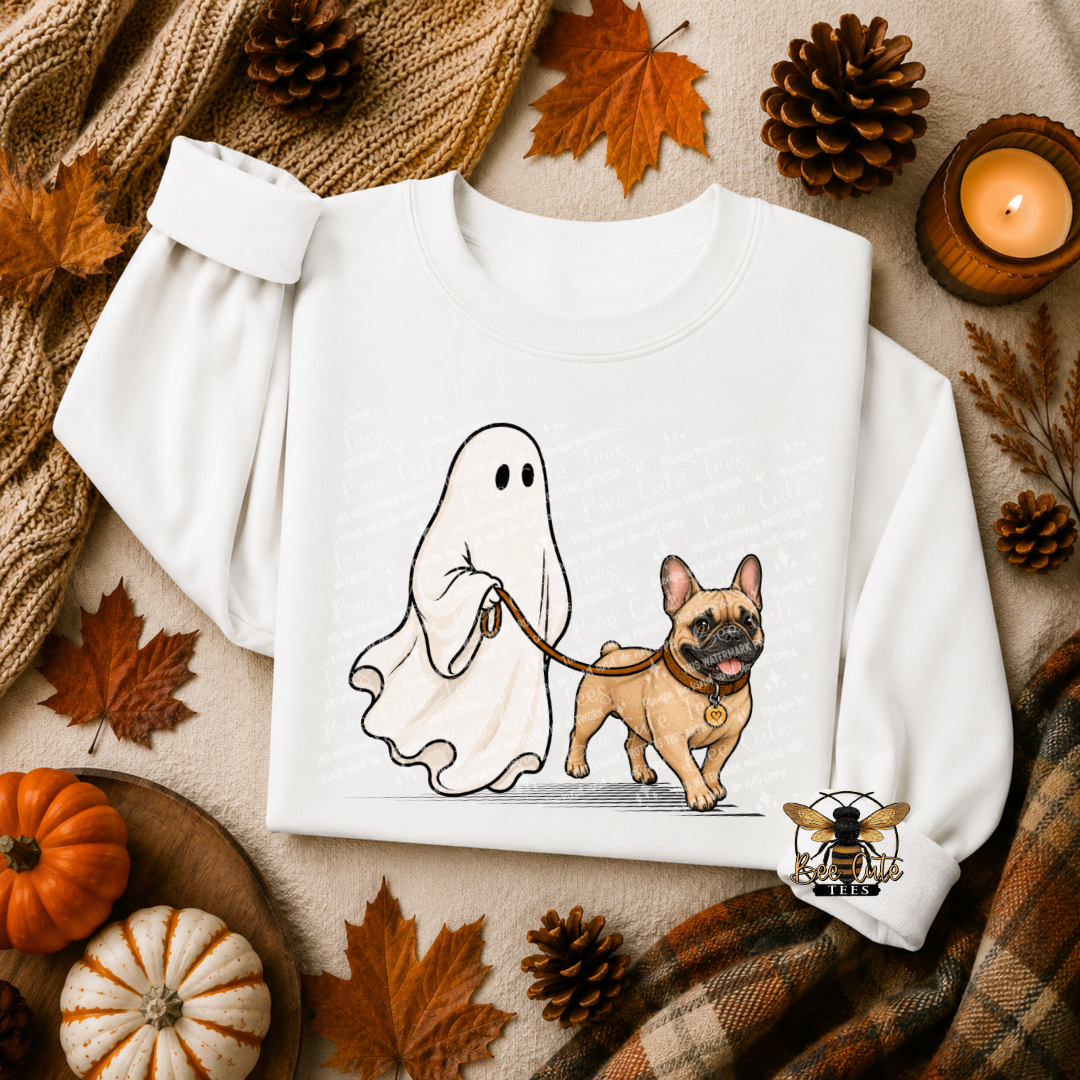 A white crewneck sweatshirt laid flat on a cozy autumn-themed background. The sweatshirt features a cartoon graphic of a ghost holding a leash next to a French Bulldog.