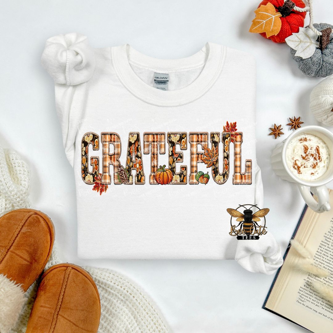 A folded white sweatshirt with a 'GRATEFUL' graphic made of autumn-themed letters, styled in a cozy flat lay with a mug of coffee, knit socks, a book, and slippers.