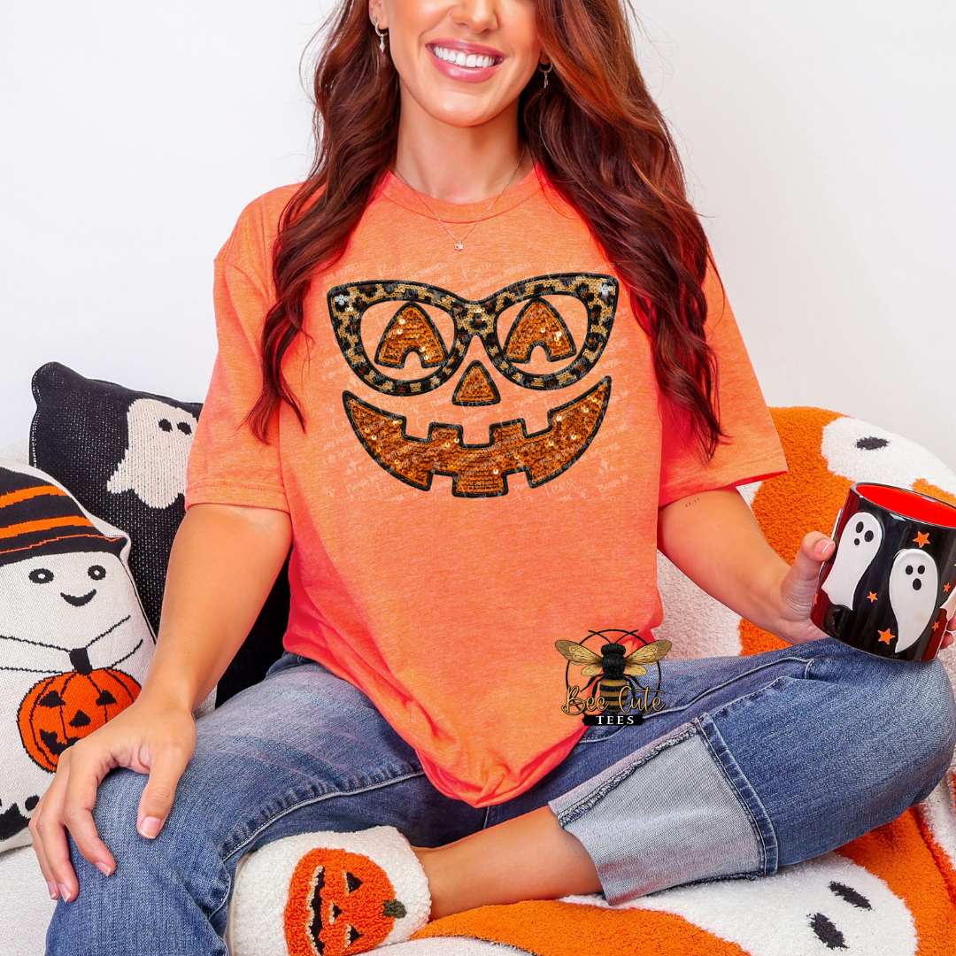 A woman wearing a bright orange short-sleeve t-shirt with a leopard-print jack-o'-lantern graphic, sitting on a cozy white and orange couch and holding a black penguin mug.