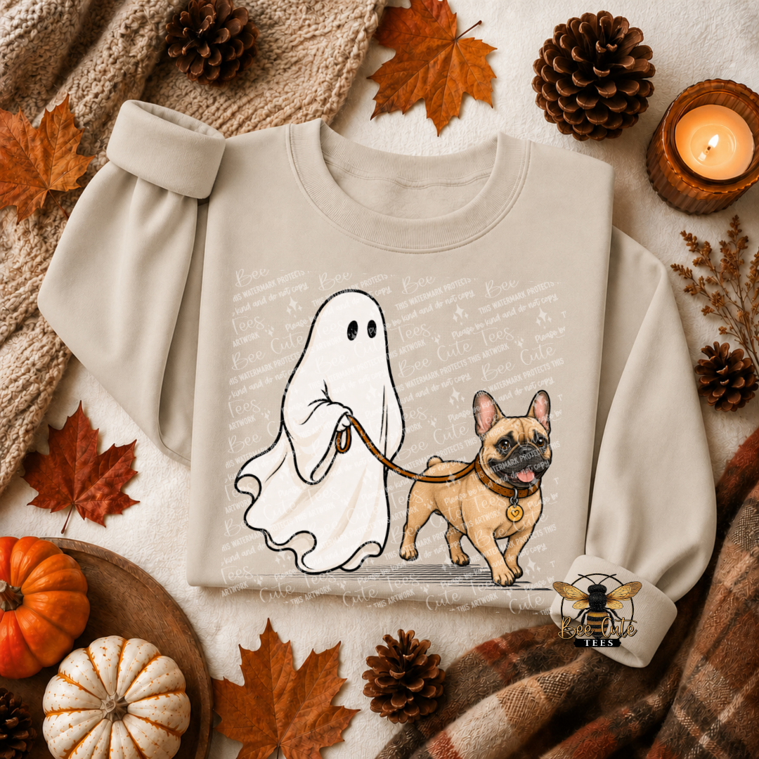 A beige crewneck sweatshirt featuring a cartoon graphic of a white ghost on a leash walking a tan French Bulldog. The sweatshirt is styled in an autumn flat-lay setting with a knit blanket, pinecones, maple leaves, small pumpkins, and a lit candle.