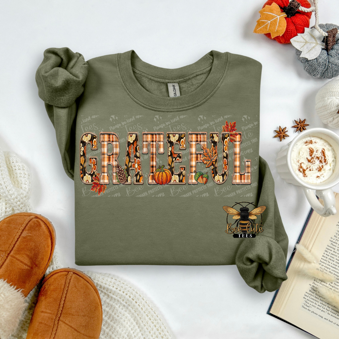 An olive green long-sleeve t-shirt with a 'GRATEFUL' autumn-themed graphic, styled in a cozy flat-lay arrangement with brown slippers, a mug of coffee, an open book, and fall decorations.