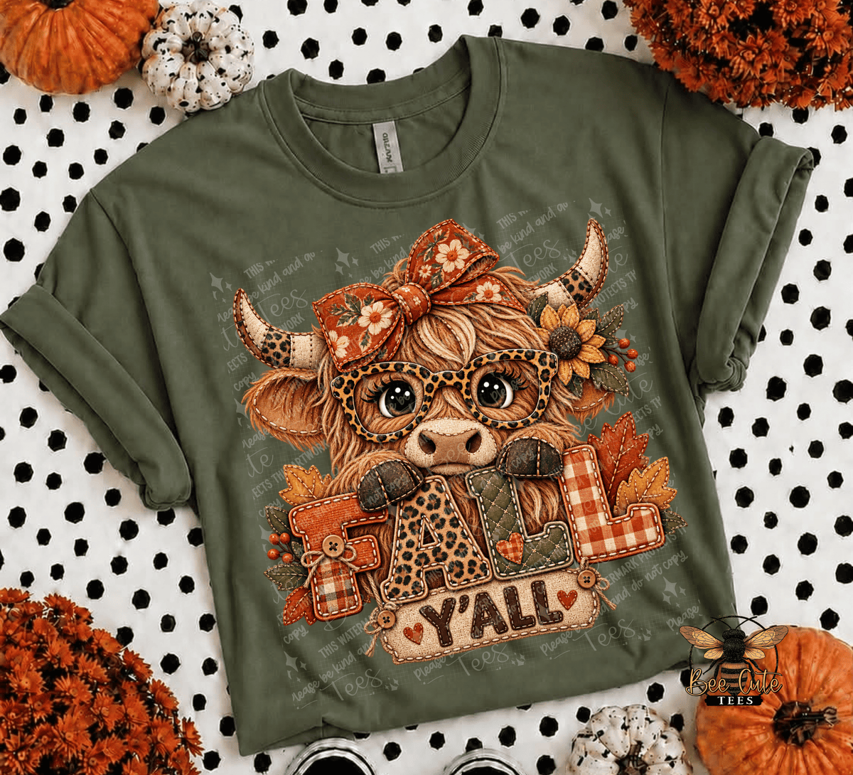 An olive green short-sleeve t-shirt with a cartoon Highland cow graphic holding the word 'Y'all' in autumn-themed patches, laid flat on a black and white polka dot background with pumpkins and fall decorations.