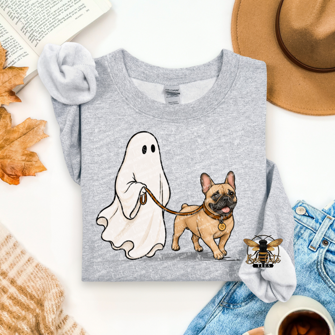 A folded heather gray graphic T-shirt is styled in a flat lay with a ghost and a French bulldog illustration on the front. The shirt is surrounded by seasonal props including autumn leaves, an open book, a tan hat, blue jeans, a knit sweater, a bee hair clip, and a cup of coffee on a white surface.