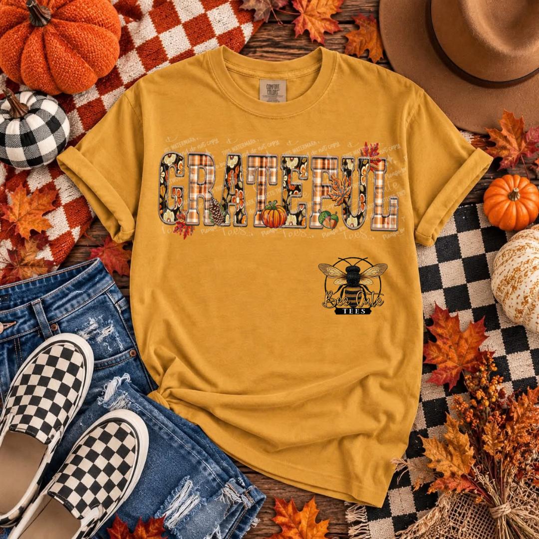 A mustard yellow graphic t-shirt laid flat in an autumn-themed flat lay with jeans, checkered shoes, pumpkins, fall leaves, a hat, and a plaid blanket. The shirt features the word 'GRATEFUL' in a decorative plaid pattern with small illustrations, and a bee logo below.