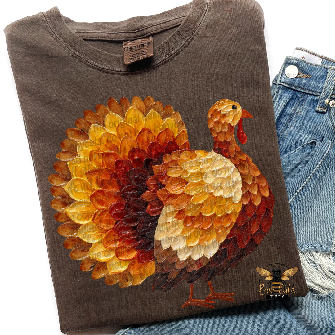 A folded dark brown t-shirt with a large, colorful painted turkey graphic on the front, styled next to a pair of blue denim shorts. 