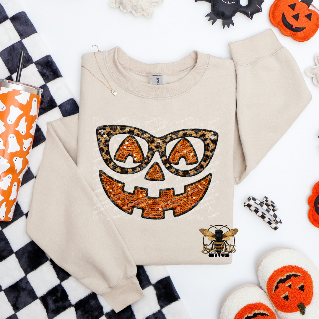 A beige crewneck sweatshirt with a Halloween-themed graphic of a jack-o'-lantern face wearing leopard-print sunglasses, styled in a flat lay with seasonal props like a checkered blanket, a ghost-patterned tumbler, and pumpkin decorations.
