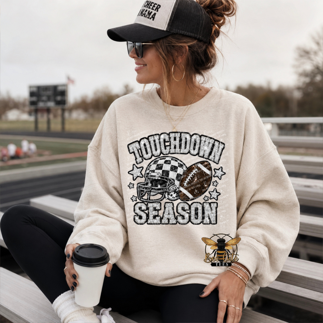 A woman sitting on outdoor bleachers wearing a cream-colored sweatshirt with a distressed 'TOUCHDOWN SEASON' football graphic on the front. She is also wearing a black and white cheerleader cap, sunglasses, and black leggings, holding a coffee cup.