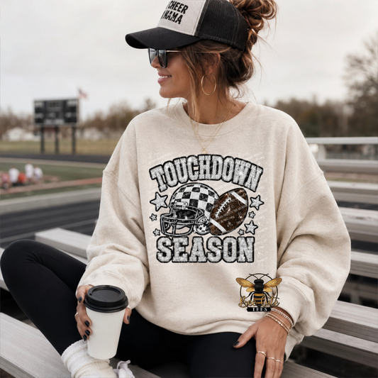 A woman sitting on outdoor bleachers wearing a cream-colored sweatshirt with a distressed 'TOUCHDOWN SEASON' football graphic on the front. She is also wearing a black and white cheerleader cap, sunglasses, and black leggings, holding a coffee cup.