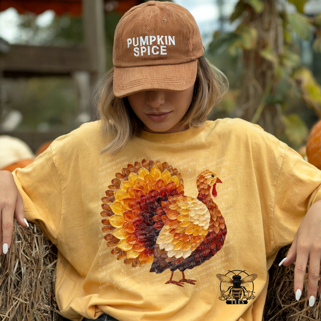 A woman wearing a tan 'Pumpkin Spice' baseball cap and a yellow graphic t-shirt with a large turkey design, sitting on a hay bale in an outdoor autumn setting.
