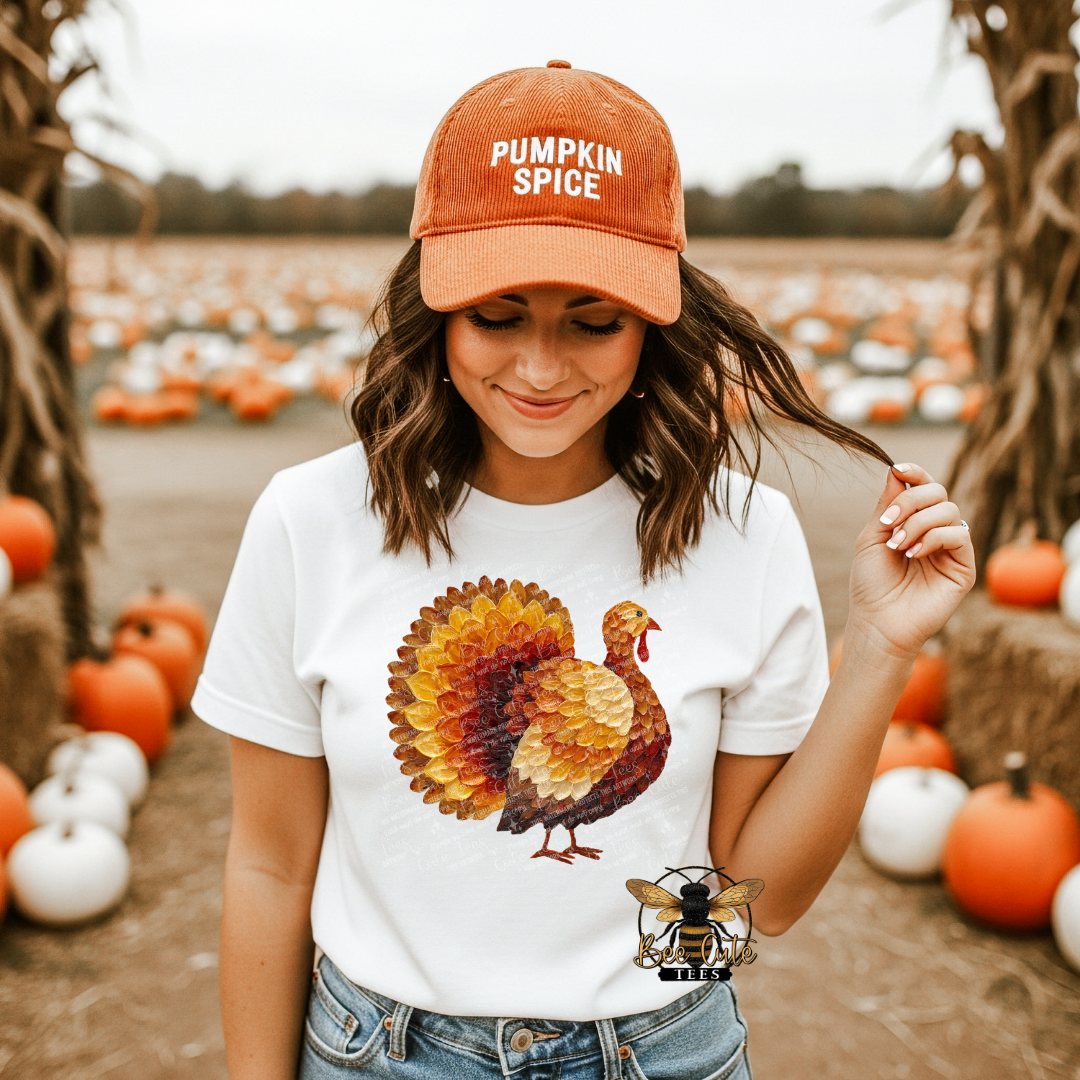 A woman standing in a pumpkin patch wearing a rust-orange baseball cap that says 'PUMPKIN SPICE' and a white graphic T-shirt featuring a large autumn turkey design. She is also wearing blue jeans, with pumpkins and cornstalks visible in the background.