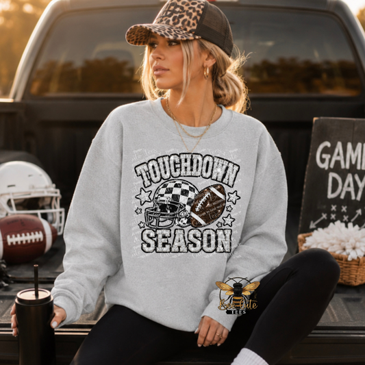 A woman sitting in the bed of a pickup truck wearing a light gray sweatshirt with a distressed 'TOUCHDOWN SEASON' football graphic. The graphic features a helmet with a checkered pattern, a football, and stars.