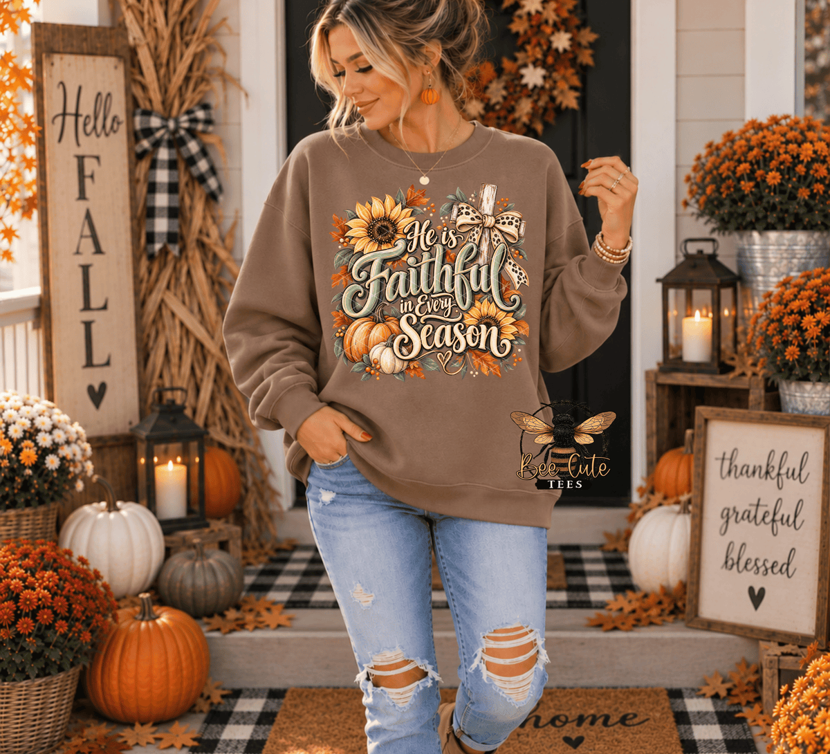 A woman stands on a decorated autumn porch wearing a taupe sweatshirt with a colorful 'He is faithful in every season' graphic featuring sunflowers, pumpkins, and a bee. She is paired with distressed blue jeans.