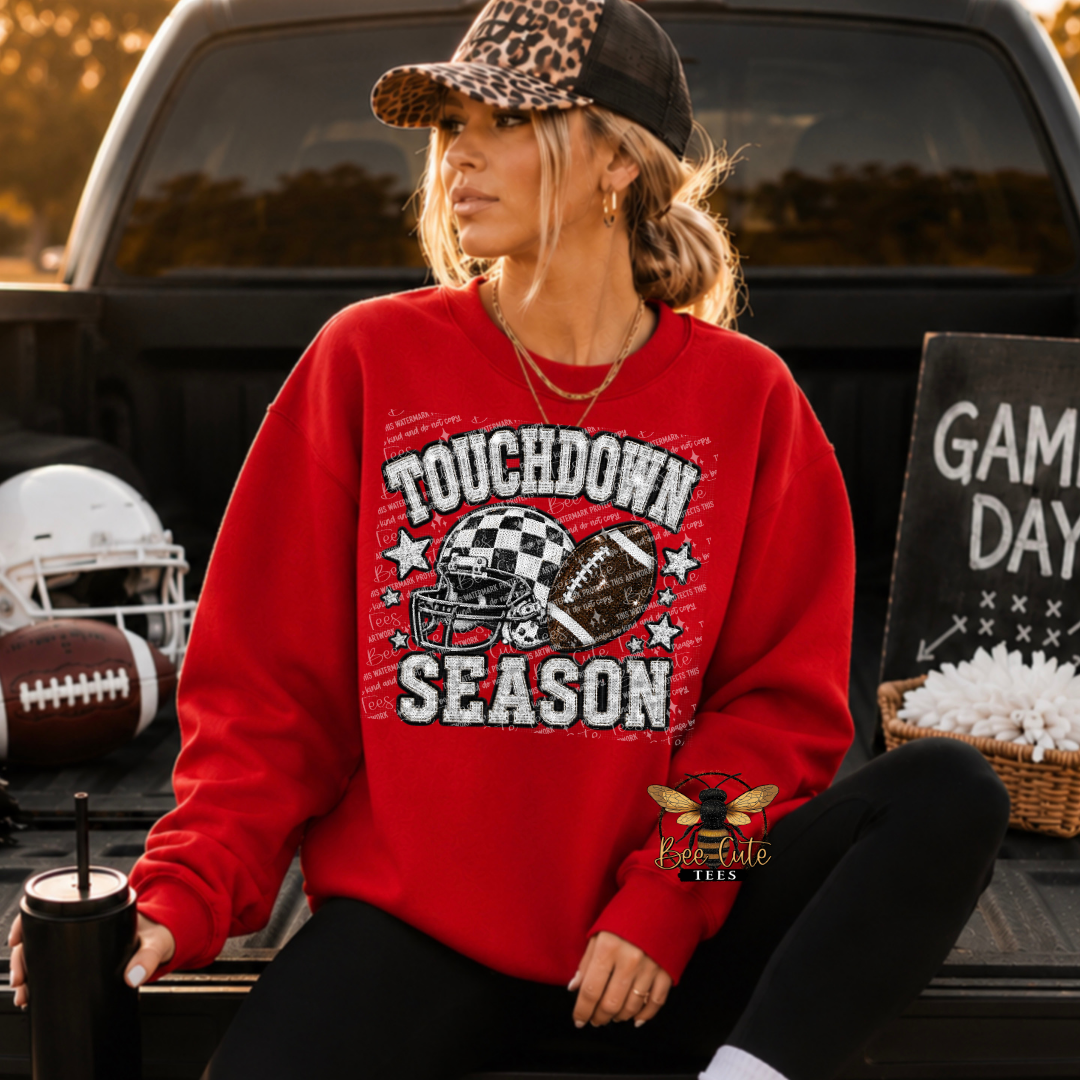 A woman wearing a red graphic sweatshirt with 'TOUCHDOWN SEASON' and football helmets sits in the bed of a pickup truck. The sweatshirt features a distressed white graphic with black outlines.