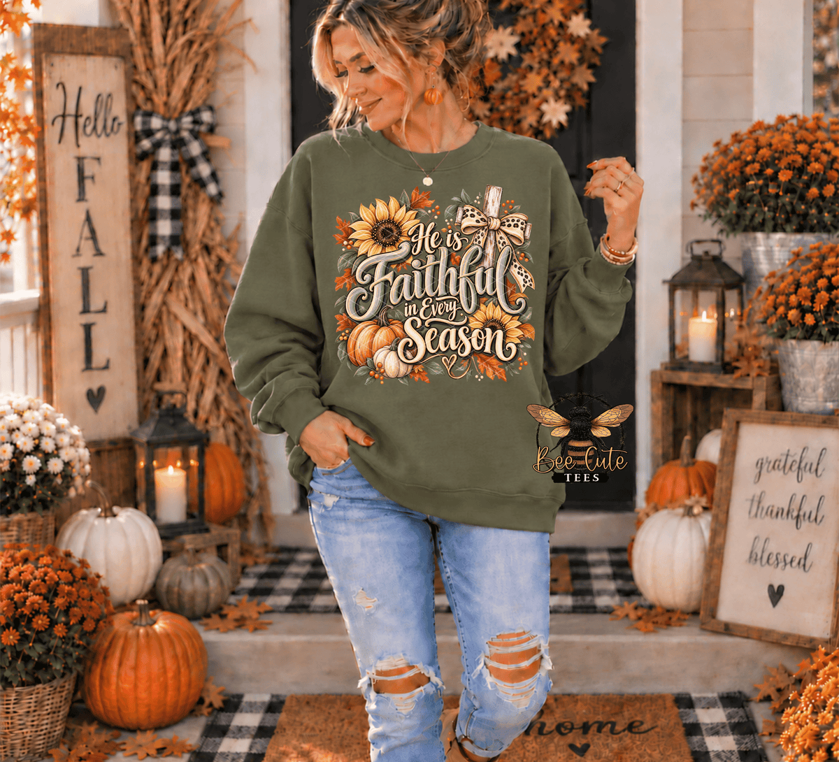 A woman stands on a decorated autumn front porch wearing an olive green sweatshirt with a large fall-themed graphic that reads 'He is faithful in every season' with sunflowers, pumpkins, and leaves. She is also wearing distressed light blue jeans.