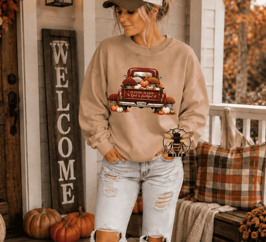 A woman wearing a beige crewneck sweatshirt with a fall harvest graphic of a red truck and pumpkins, paired with distressed light-wash jeans, standing on a decorated autumn porch.