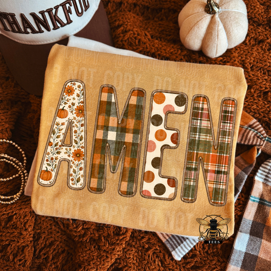 A folded mustard yellow folded t-shirt, laid on a brown shag rug with autumn-themed props. The cloth features the word “AMEN” in large decorative letters: A and M have floral vine patterns, N has orange polka dots on cream, and E has an orange-brown plaid design.