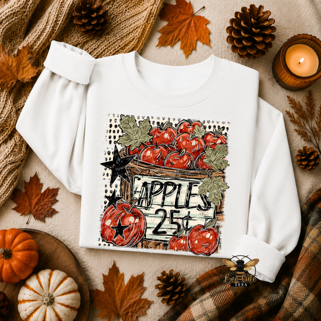 A white long-sleeve crewneck sweatshirt laid flat on a beige fabric background with autumn props including pinecones, leaves, pumpkins, a knitted scarf, and a lit candle. The front has a large fall-themed apple graphic with the text 'APPLES 25¢' and a bee watermark near the lower right.