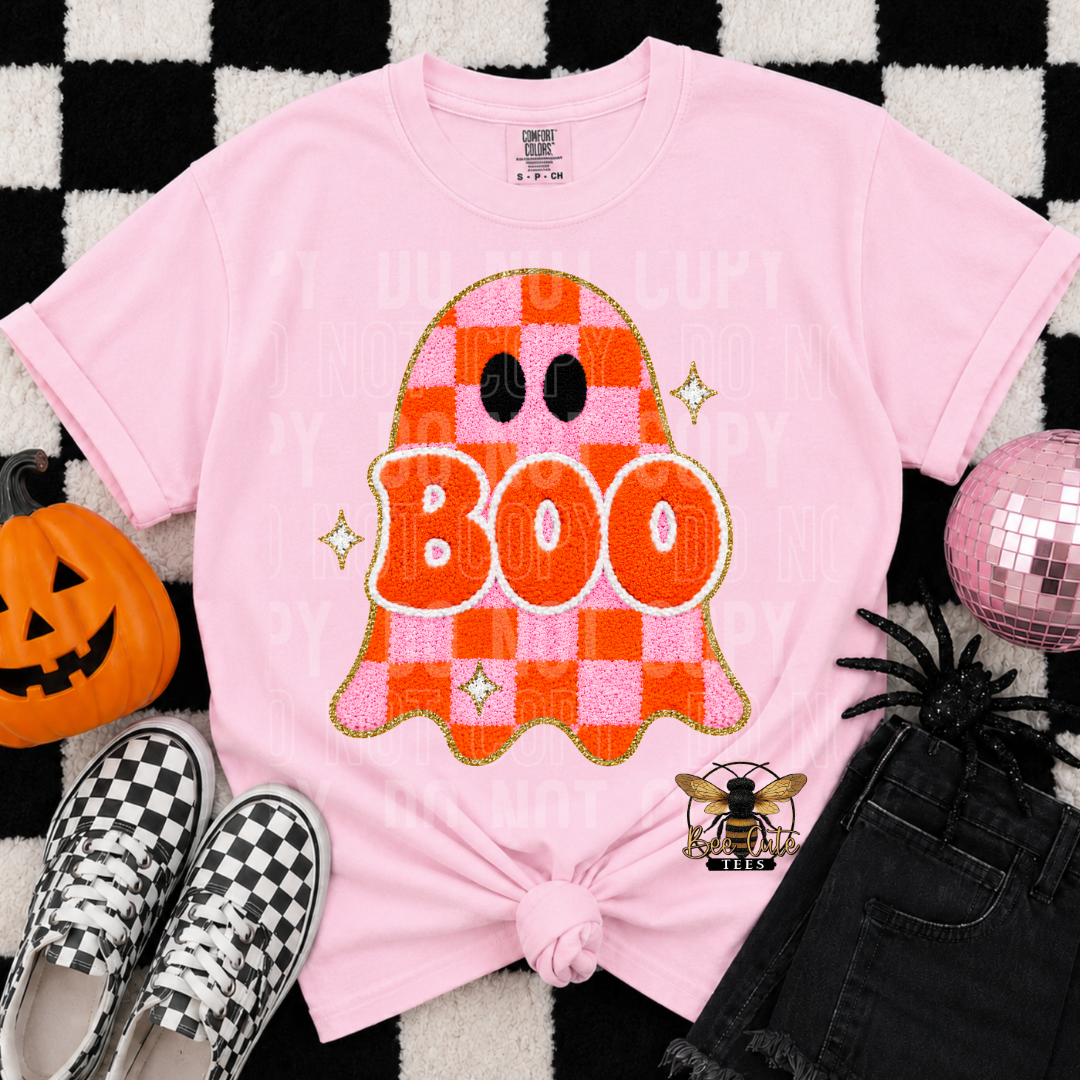 A pink short-sleeve graphic T-shirt laid flat on a black-and-white checkered rug with Halloween props. The shirt has a large ghost graphic with the word 'BOO' in orange and pink checkered sections, plus small sparkle accents.