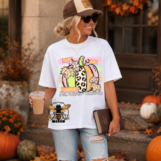 A woman wearing a white graphic t-shirt with a colorful autumn pumpkin and bee logo, paired with distressed light-wash jeans, a brown and tan baseball cap, and sunglasses. She is holding an iced coffee and a book, standing in an outdoor fall yard decorated with pumpkins and autumn foliage.